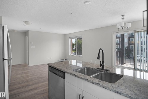 Kitchen island featuring a double basin stainless steel sink with a matte black faucet, granite-finish countertop, and integrated stainless steel dishwasher - 202 142 Ebbers Boulevard, Edmonton, AB 