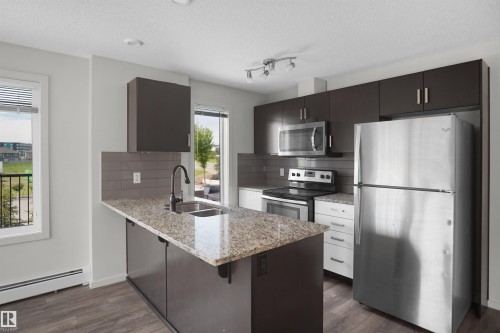 Modern kitchen featuring an island with a double basin sink, stainless steel appliances, dark wood-finish cabinetry, and a tile backsplash - 202 142 Ebbers Boulevard, Edmonton, AB 
