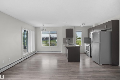 Open-concept living area featuring wood-finish flooring, a modern kitchen island with an undermount sink, stainless steel appliances, and dark cabinetry - 202 142 Ebbers Boulevard, Edmonton, AB 