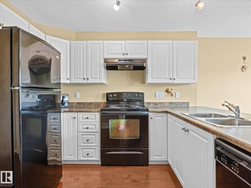 Kitchen featuring white cabinetry, dark countertops, a black electric range, a black refrigerator, and wood-finish flooring - #88 5001 62 St, Beaumont, AB - Indoor Photo Showing Kitchen With Double Sink