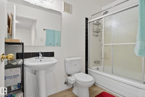 Pedestal sink with black tile backsplash, full vanity mirror, built-in tub with sliding glass shower door, and wood-finish flooring - 204 12404 114 Avenue, Edmonton, AB - Indoor Photo Showing Bathroom