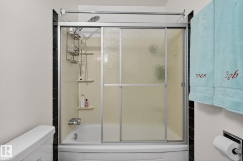 Bathtub and shower combination featuring sliding textured glass doors, black tiled surround, and white wall panels - 204 12404 114 Avenue, Edmonton, AB - Indoor Photo Showing Bathroom
