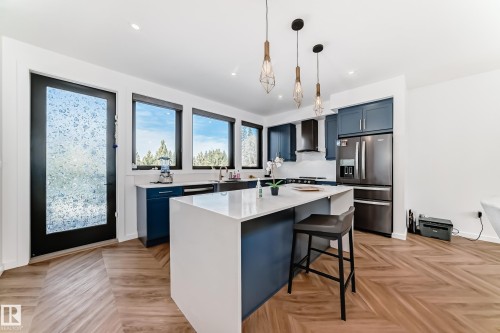 Modern kitchen featuring a large central island with a white countertop, blue cabinetry, stainless steel appliances, and wood-finish flooring with a herringbone pattern - 46 1304 Rutherford Road, Edmonton, AB - Indoor Photo Showing Kitchen With Upgraded Kitchen