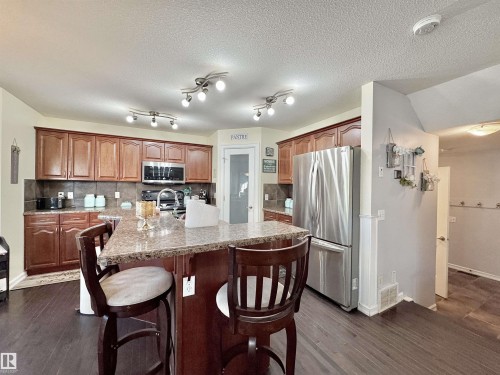 Kitchen featuring wood-finish flooring, an island with a stone-finish countertop, wood cabinetry, stainless steel appliances, and track lighting - 105 Greystone Crescent, Spruce Grove, AB 
