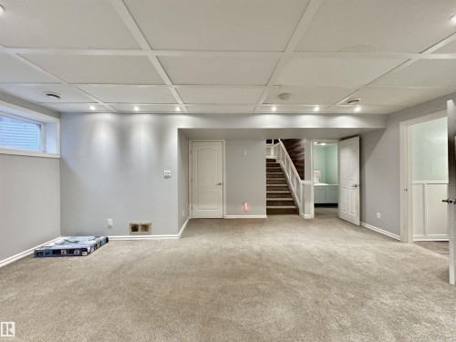 Spacious finished room featuring neutral carpet flooring, recessed lighting, a coffered ceiling, and a visible staircase with carpeted treads - 105 Greystone Crescent, Spruce Grove, AB 