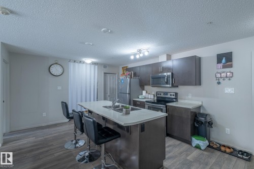 Kitchen featuring an island with a light-toned countertop and dual basin sink, dark wood-finish cabinetry, stainless steel appliances, track lighting, and wood-finish flooring - 107 142 Ebbers Boulevard, Edmonton, AB 