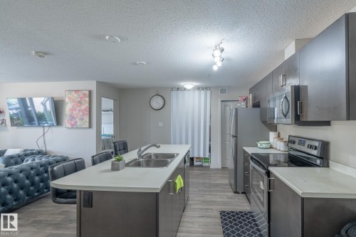 Open-concept kitchen featuring an island with a double basin sink, dark cabinetry, light countertops, and wood-finish flooring - 107 142 Ebbers Boulevard, Edmonton, AB 