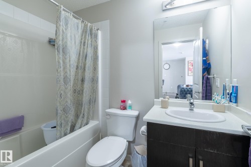 Bathroom featuring a white bathtub, white tiled shower surround, dark wood-finish vanity, white countertop, and rectangular mirror - 107 142 Ebbers Boulevard, Edmonton, AB 