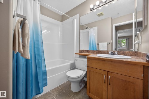 Bathroom featuring a wood-finish vanity with a built-in sink, a large mirror, a four-bulb vanity light fixture, a white bathtub with shower surround, and tile flooring - 111 3425 19, Edmonton, AB 
