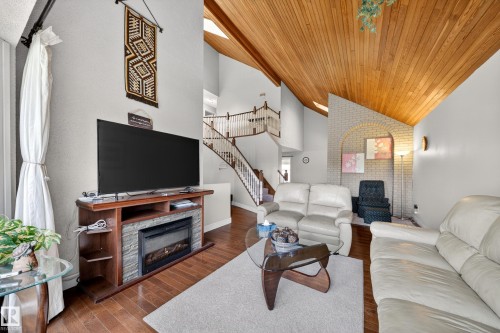 Vaulted wood-plank ceilings complement the living area featuring wood-finish flooring, a built-in electric fireplace with stone-look surround, and a two-story staircase with wood railings - 34 Grand Meadow Crescent, Edmonton, AB 