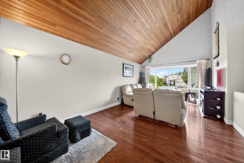 Spacious living area featuring a vaulted ceiling with wood-finish paneling, rich wood-finish flooring, a broad picture window, and neutral wall tones - 34 Grand Meadow Crescent, Edmonton, AB 