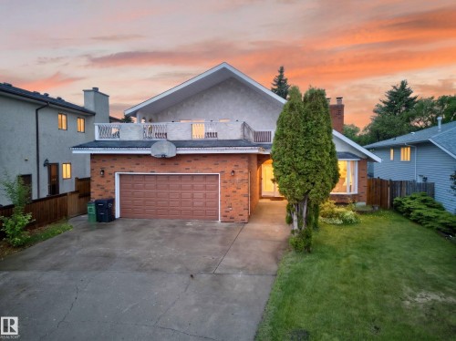 Brick and stucco exterior featuring a two-car garage with a brown door, a front lawn with mature landscaping, and a bay window - 34 Grand Meadow Crescent, Edmonton, AB 