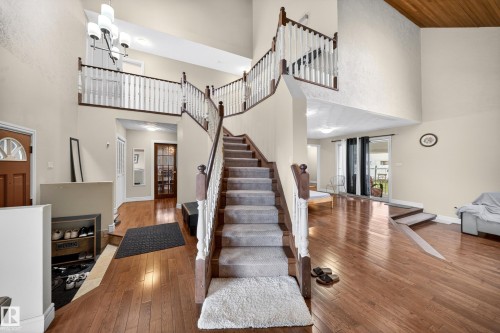 Grand entryway featuring a double staircase with wood railings and white spindles, wood-finish flooring, a vaulted ceiling with a wood-paneled accent, and a large multi-light chandelier - 34 Grand Meadow Crescent, Edmonton, AB 