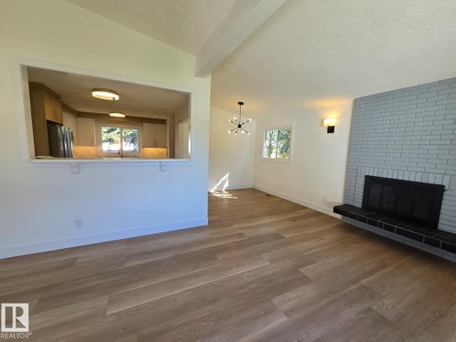 Spacious living area featuring wood-finish flooring, a painted brick fireplace with a tiled hearth, and a pass-through opening to the kitchen - 18526 86A Avenue, Edmonton, AB - Indoor With Fireplace