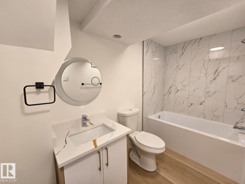 Bathroom featuring a white vanity with gold-tone hardware, a white countertop with integrated sink, a circular back-lit mirror, a white toilet, and a white bathtub with white and gray patterned wall tiling - 18526 86A Avenue, Edmonton, AB - Indoor Photo Showing Bathroom
