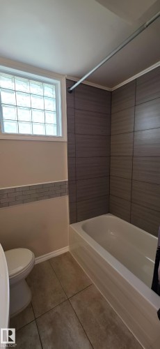 Contemporary bathroom featuring a white bathtub, large format tiled shower surround, block glass window, and coordinated floor tiling - 6704 93 Street, Edmonton, AB 
