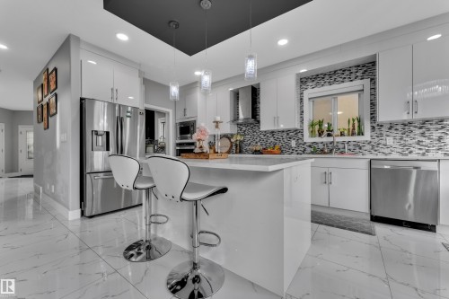 Modern kitchen featuring a central island with a light-toned countertop, glossy white cabinetry, stainless steel appliances, a mosaic tile backsplash, and large format marble-look floor tiling - 2419 15 Avenue, Edmonton, AB 