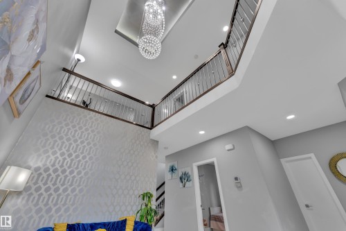 Two-story interior with a tiered crystal chandelier, upper-level landing with dark wood railing and metal spindles, recessed lighting, and a textured accent wall - 2419 15 Avenue, Edmonton, AB 