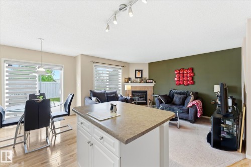 Open-concept living space featuring wood-finish flooring, a kitchen island with storage, a dining area with a glass patio door, a living room with carpet flooring, and a fireplace with a mantel - 37 4020 21 Street, Edmonton, AB 