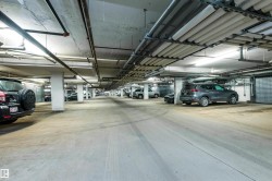 Underground parking garage featuring concrete flooring, structural support columns, overhead fluorescent lighting, exposed utility piping, and individual storage units - 