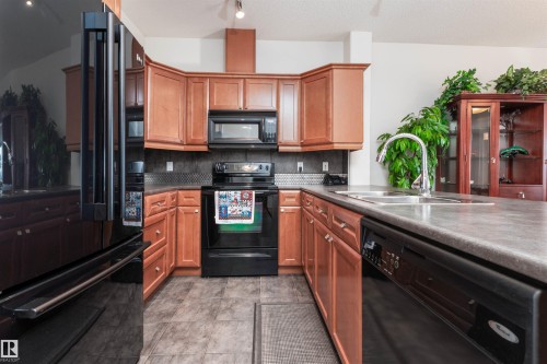 U-shaped kitchen featuring wood cabinetry, dark countertops, black appliances, a double basin sink with a high-arc faucet, and tiled flooring - 439 300 Palisades Way, Sherwood Park, AB - Indoor Photo Showing Kitchen With Double Sink