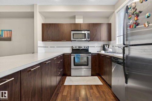U-shaped kitchen featuring wood-finish cabinetry, stainless steel appliances, light-toned countertops, a white subway tile backsplash, and wood-finish flooring - 30 6075 Schonsee Way, Edmonton, AB 