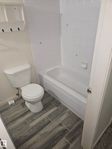 Bathroom featuring a white toilet, white bathtub with white tiled surround, and wood-finish tile flooring - 401 10615 110 Street, Edmonton, AB 