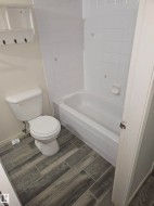 Bathroom featuring a white toilet, white bathtub with white tiled surround, and wood-finish tile flooring - 