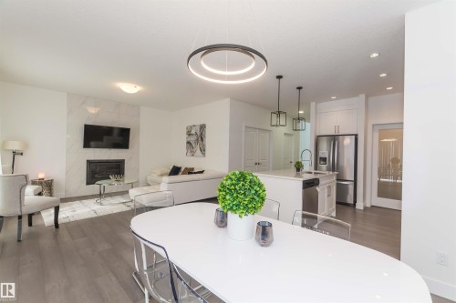 Open-concept living space featuring wood-finish flooring, a contemporary fireplace with marble-look surround, a kitchen island with pendant lighting, white cabinetry, and stainless steel appliances - 15168 16 Street Nw, Edmonton, AB 