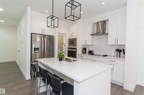 Contemporary kitchen featuring a large central island with an undermount double sink, white cabinetry with silver hardware, stainless steel appliances, a white subway tile backsplash, and recessed and pendant lighting - 15168 16 Street Nw, Edmonton, AB 