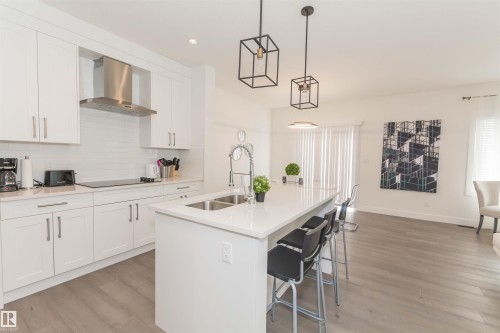 Modern kitchen featuring white cabinetry, a large center island with seating, a stainless steel range hood, white subway tile backsplash, and wood-finish flooring - 15168 16 Street Nw, Edmonton, AB 