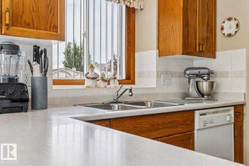 Kitchen featuring a double basin stainless steel sink, light-toned countertops, wood cabinetry, a tiled backsplash, and a built-in dishwasher - 5131 152A Avenue, Edmonton, AB 