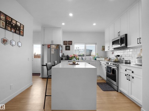 Modern kitchen featuring a large central island with seating, white shaker cabinetry, stainless steel appliances, and wood-finish flooring - 19316 29 Avenue, Edmonton, AB 