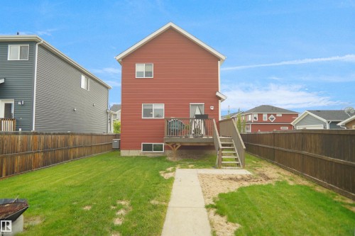Rear exterior featuring a red siding facade, elevated wood deck with stairs, a concrete path, and a fenced backyard with grass - 292 Simmonds Way, Leduc, AB 