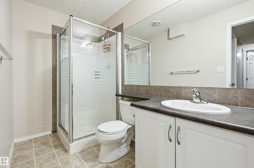 Bathroom featuring a glass-enclosed shower, white vanity cabinetry, a dark countertop, a rectangular mirror, and neutral-toned floor tiling - 1804 121 Street, Edmonton, AB 