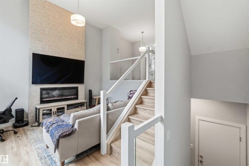 Living area featuring a textured accent wall, integrated fireplace, wood-finish flooring, and a contemporary glass panel staircase - 33 1729 Keene Crescent, Edmonton, AB 