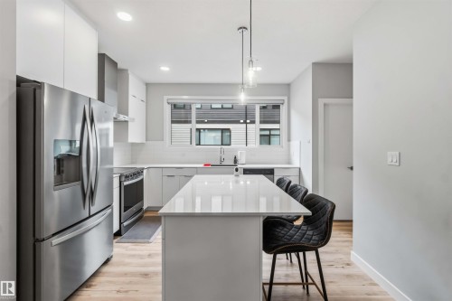 Modern kitchen featuring an expansive island with a polished countertop, stainless steel appliances, white flat-panel cabinetry, a white subway tile backsplash, and wood-finish flooring - 33 1729 Keene Crescent, Edmonton, AB 