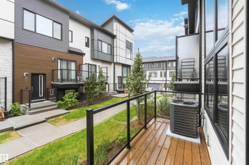 Contemporary townhome featuring mixed material exterior siding, a private wood-finish balcony with glass railings, a black entry door with frosted glass, and mature landscaping - 33 1729 Keene Crescent, Edmonton, AB 