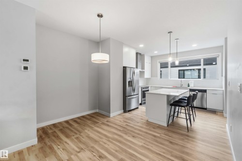 Open-concept kitchen and dining area featuring wood-finish flooring, white cabinetry, a kitchen island with seating, stainless steel appliances, and a window with a white frame - 33 1729 Keene Crescent, Edmonton, AB 