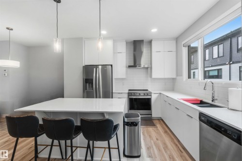 Modern kitchen featuring white cabinetry, stainless steel appliances, a large island with pendant lighting, wood-finish flooring, and a subway tile backsplash - 33 1729 Keene Crescent, Edmonton, AB 