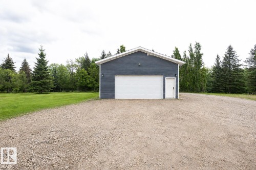 Detached garage with dark blue siding, white trim, a single overhead door, and a separate service door, situated on a gravel driveway with a grassy area and mature trees - 209 5128 Highway 18, Rural Barrhead County, AB 