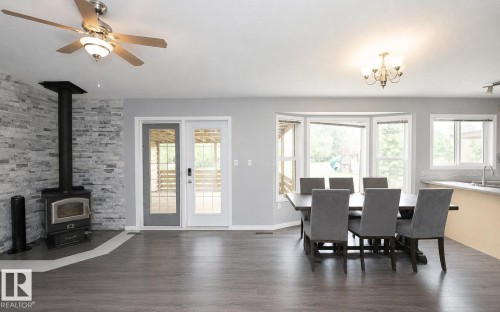 Spacious open-concept living area featuring wood-finish flooring, a stone-accented wall with a corner wood stove, and a ceiling fan - 209 5128 Highway 18, Rural Barrhead County, AB 