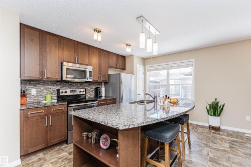 Kitchen featuring a granite-topped island with breakfast bar seating, stainless steel appliances, wood-finish cabinetry, a tiled backsplash, and recessed lighting - 31 4029 Orchards Drive, Edmonton, AB 