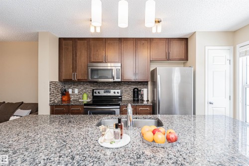 Kitchen featuring granite countertops, wood cabinetry, stainless steel appliances, and a mosaic tile backsplash - 31 4029 Orchards Drive, Edmonton, AB 