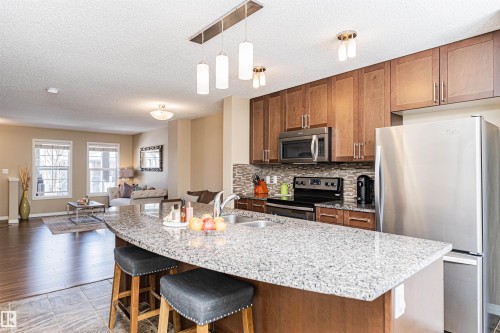 Kitchen featuring a granite-finish island with integrated sink, stainless steel appliances, wood-finish cabinetry, and a mosaic tile backsplash - 31 4029 Orchards Drive, Edmonton, AB 