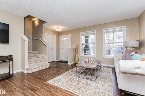 Open concept living space featuring wood-finish flooring, two large windows, a carpeted staircase with white railings, and a front entry door with transom windows - 31 4029 Orchards Drive, Edmonton, AB 
