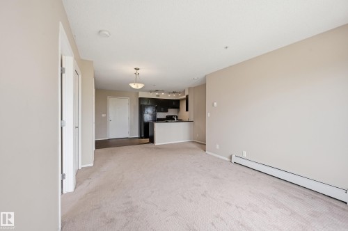 Open-concept living area with neutral carpeting, leading to a kitchen featuring dark cabinetry and black appliances - 304 11804 22 Avenue, Edmonton, AB 