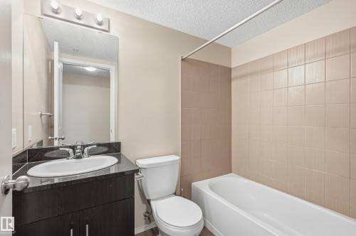 Bathroom featuring a dark wood-finish vanity with a built-in sink and dark countertop, a wall-mounted mirror with overhead lighting, a white toilet, and a bathtub with light-colored tile surround - 304 11804 22 Avenue, Edmonton, AB 