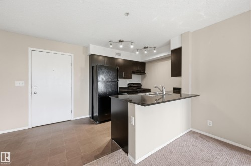 Kitchen featuring dark cabinetry, black appliances, a double basin stainless steel sink, and a breakfast bar with a dark countertop - 304 11804 22 Avenue, Edmonton, AB 
