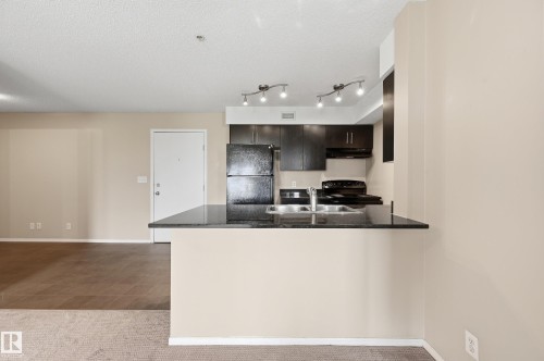 Kitchen featuring dark wood cabinetry, black appliances, track lighting, and a black countertop with an undermount sink - 304 11804 22 Avenue, Edmonton, AB 